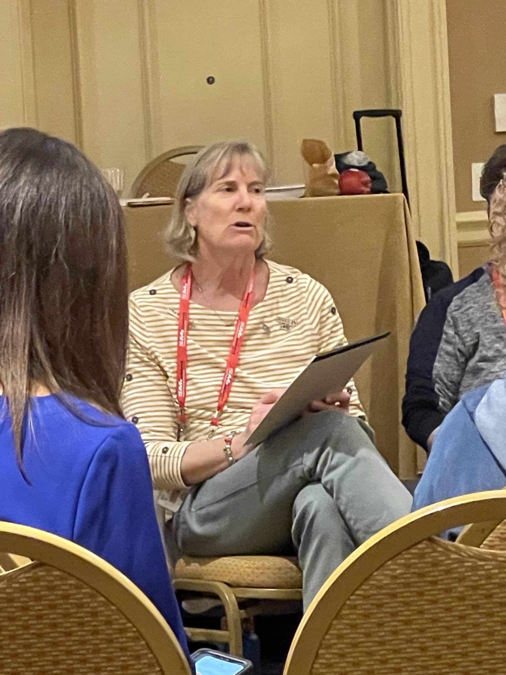 Two women seated in conversation at a conference, both wearing event lanyards.