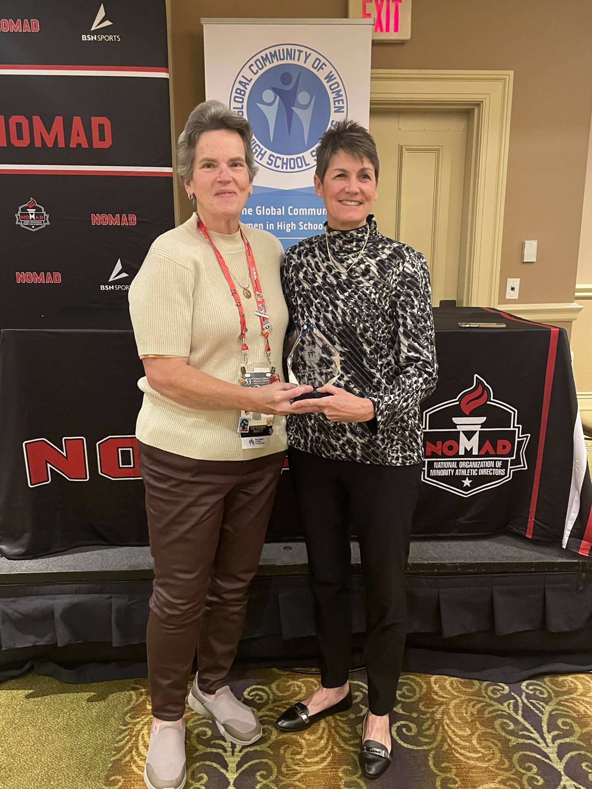 Two women at an award presentation, standing beside a Global Community of Women in High School Sports banner.