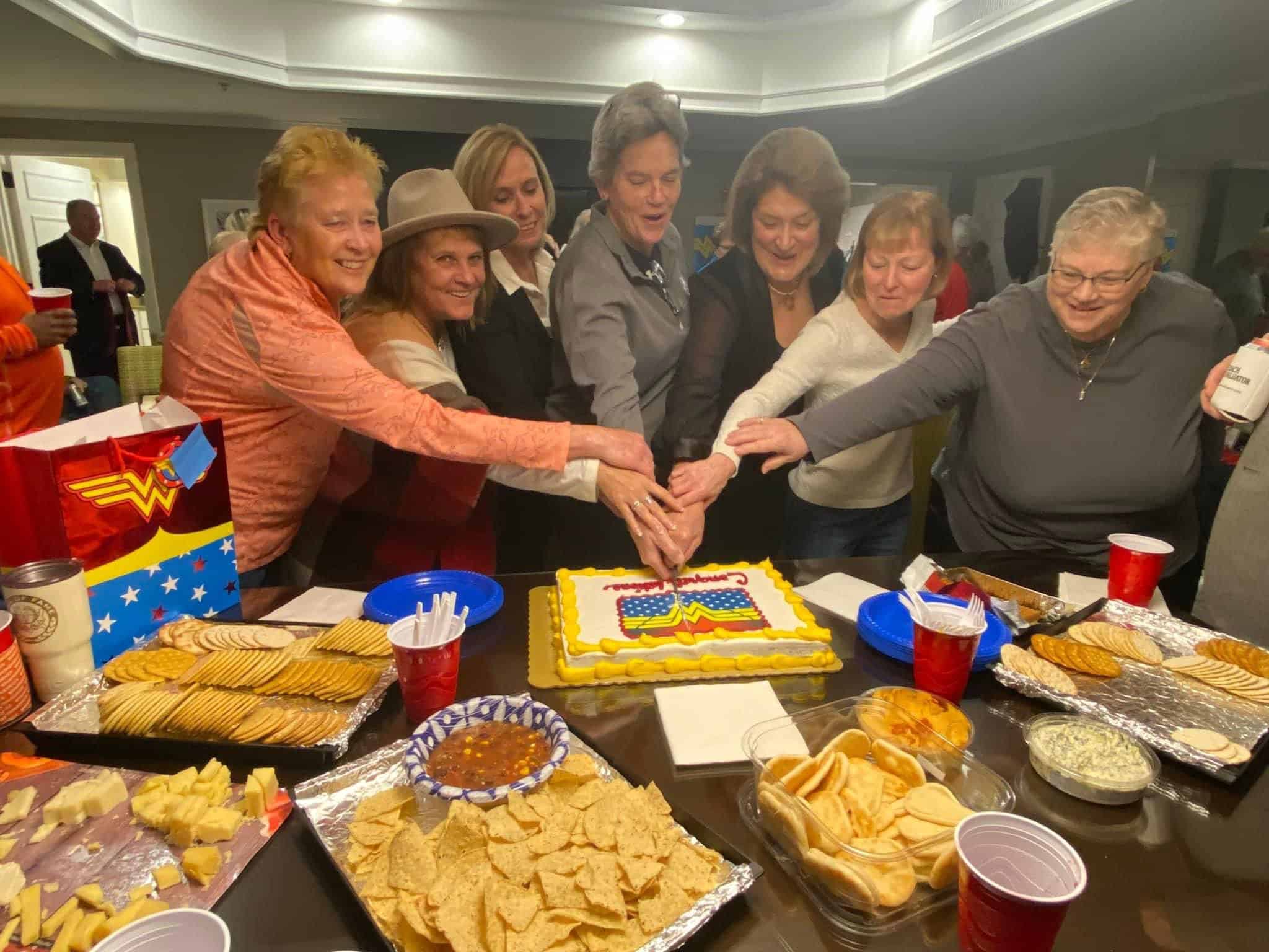 A group of women gathered around a table, cutting a celebration cake together.