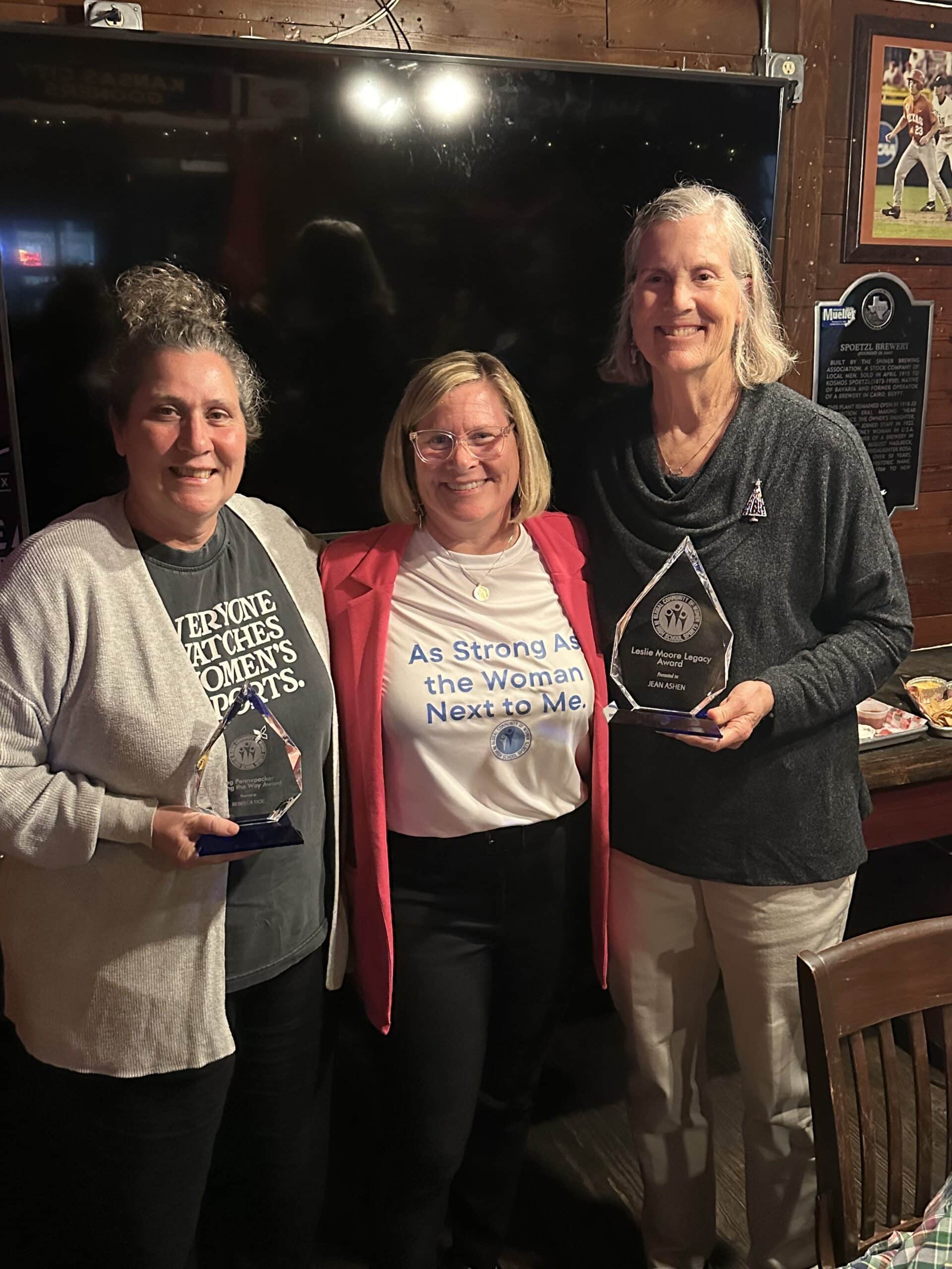 Three women holding awards together after a recognition event, one wearing a shirt that reads "As Strong As the Woman Next to Me."